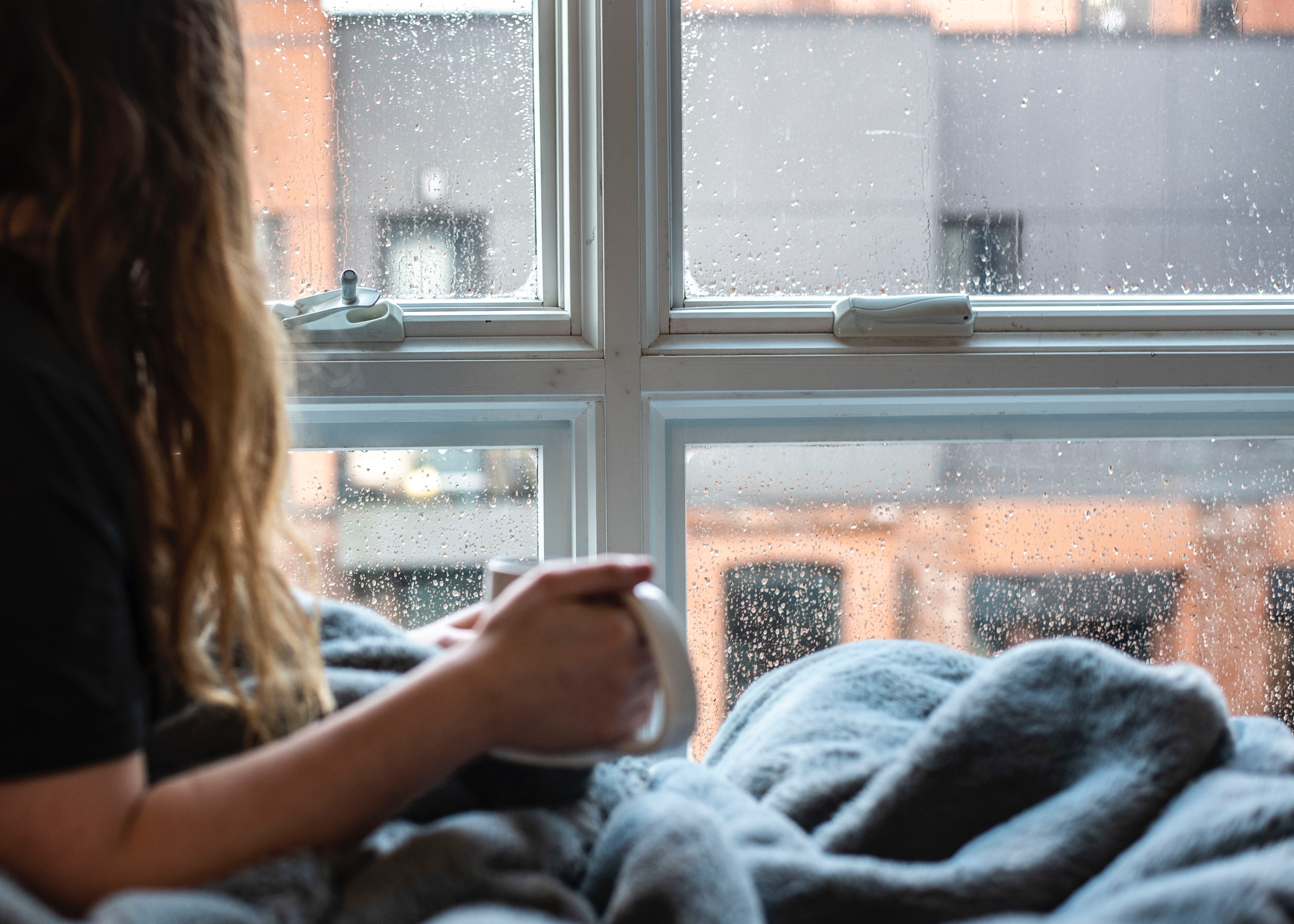 Girl sitting in bed with a mug watching cold rain outside